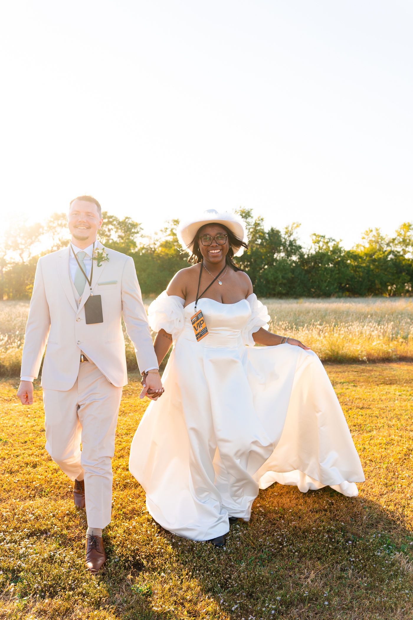 A couple in wedding attire walks through a sun-drenched field at golden hour, their white outfits glowing in the light.