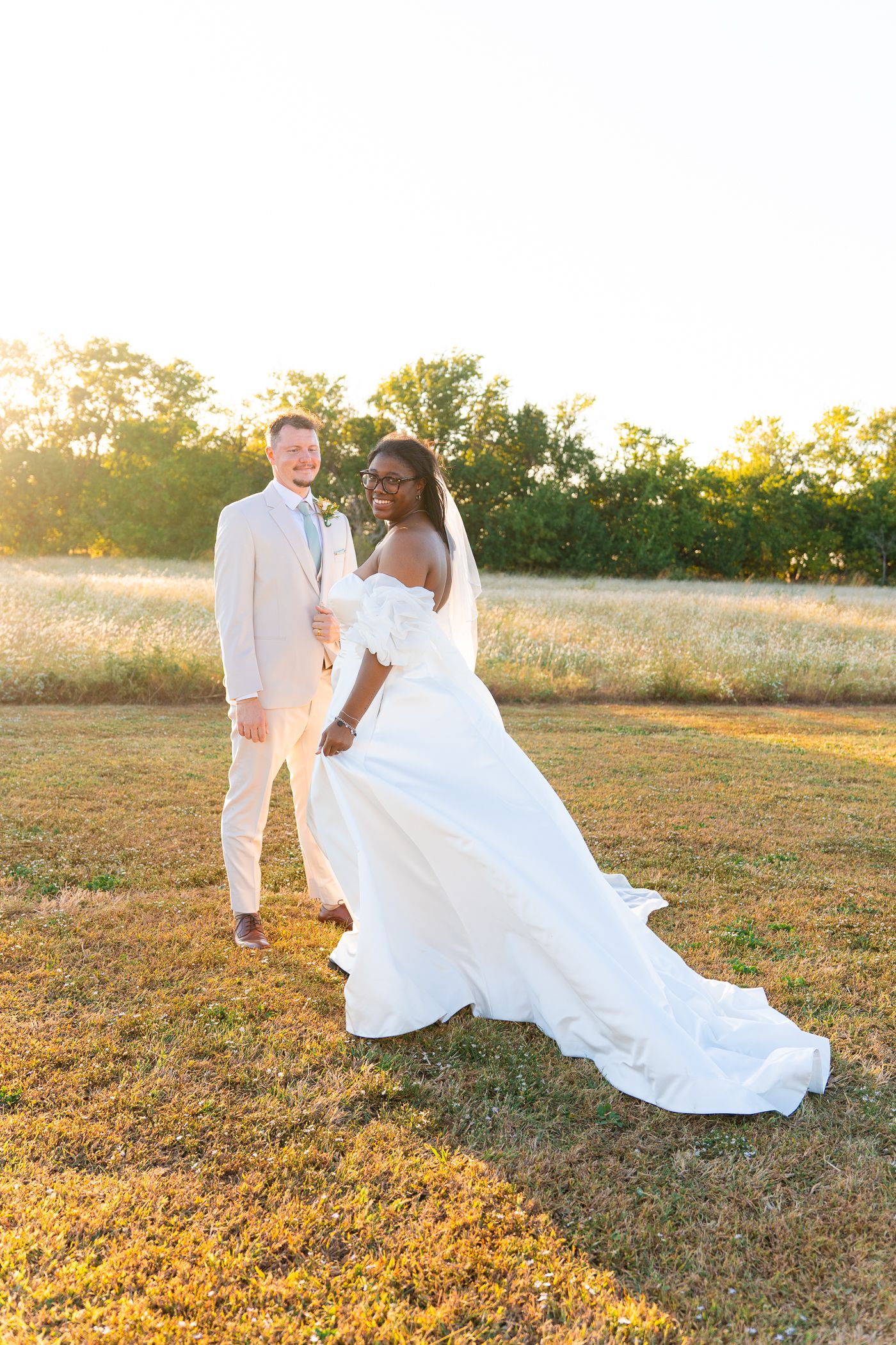 A couple in white attire walks through a golden sunlit field during sunset.