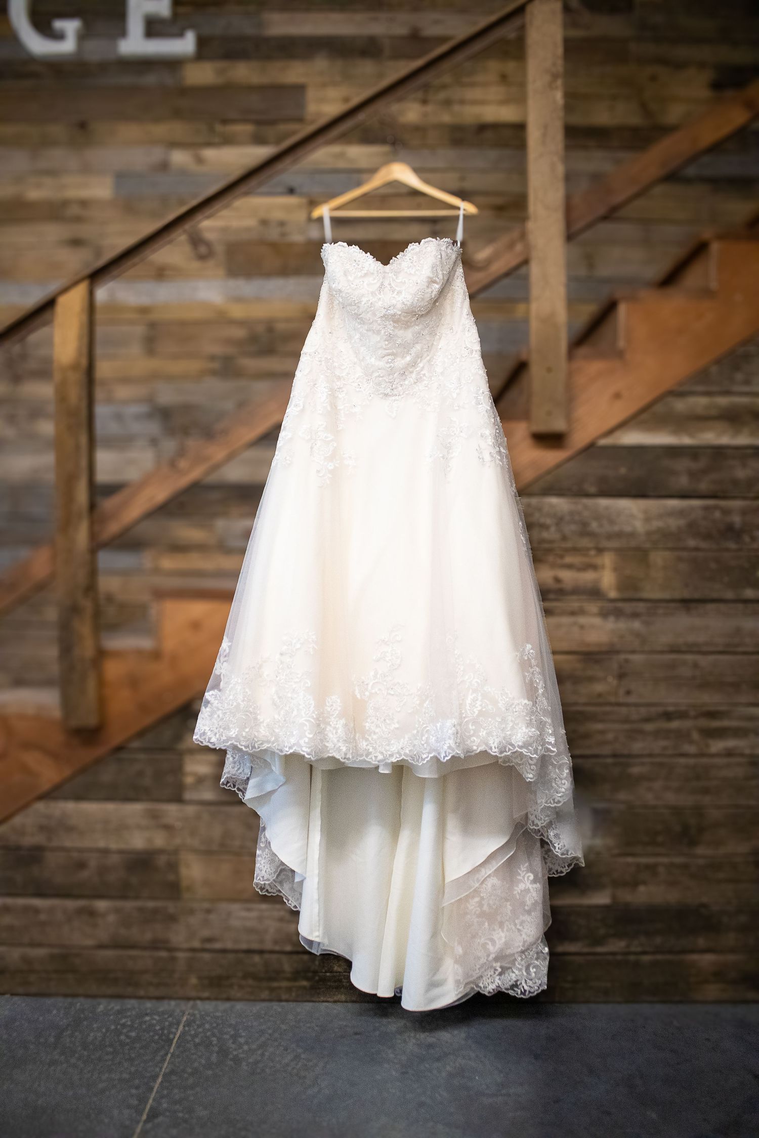 White lace wedding dress hanging against rustic wooden wall backdrop.