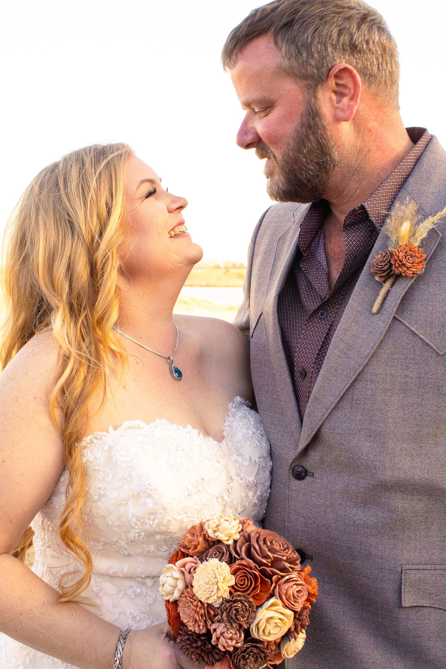 Bride in white lace dress holds autumn-colored bouquet next to groom in grey suit.