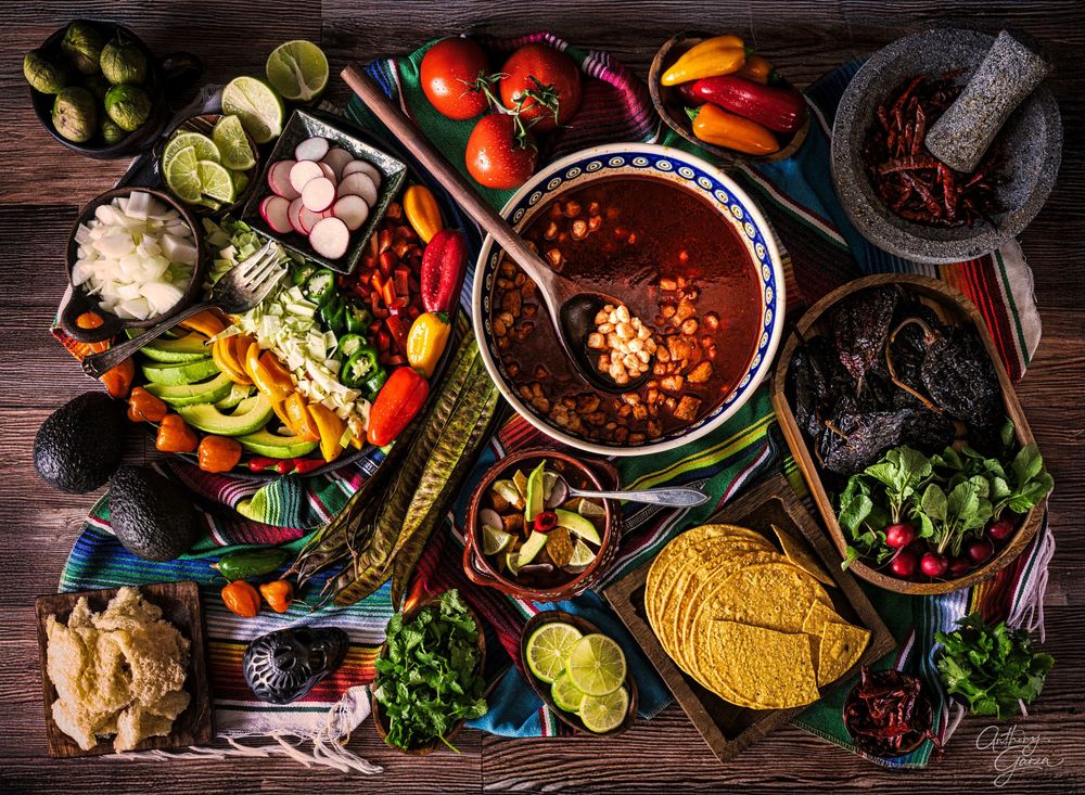 Rustic wooden table with Mexican food spread including tacos, salsas, limes, peppers and fresh ingredients.
