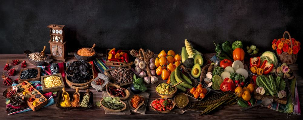 A vibrant display of fresh produce, grilled foods, and prepared dishes arranged in a dark rustic setting.