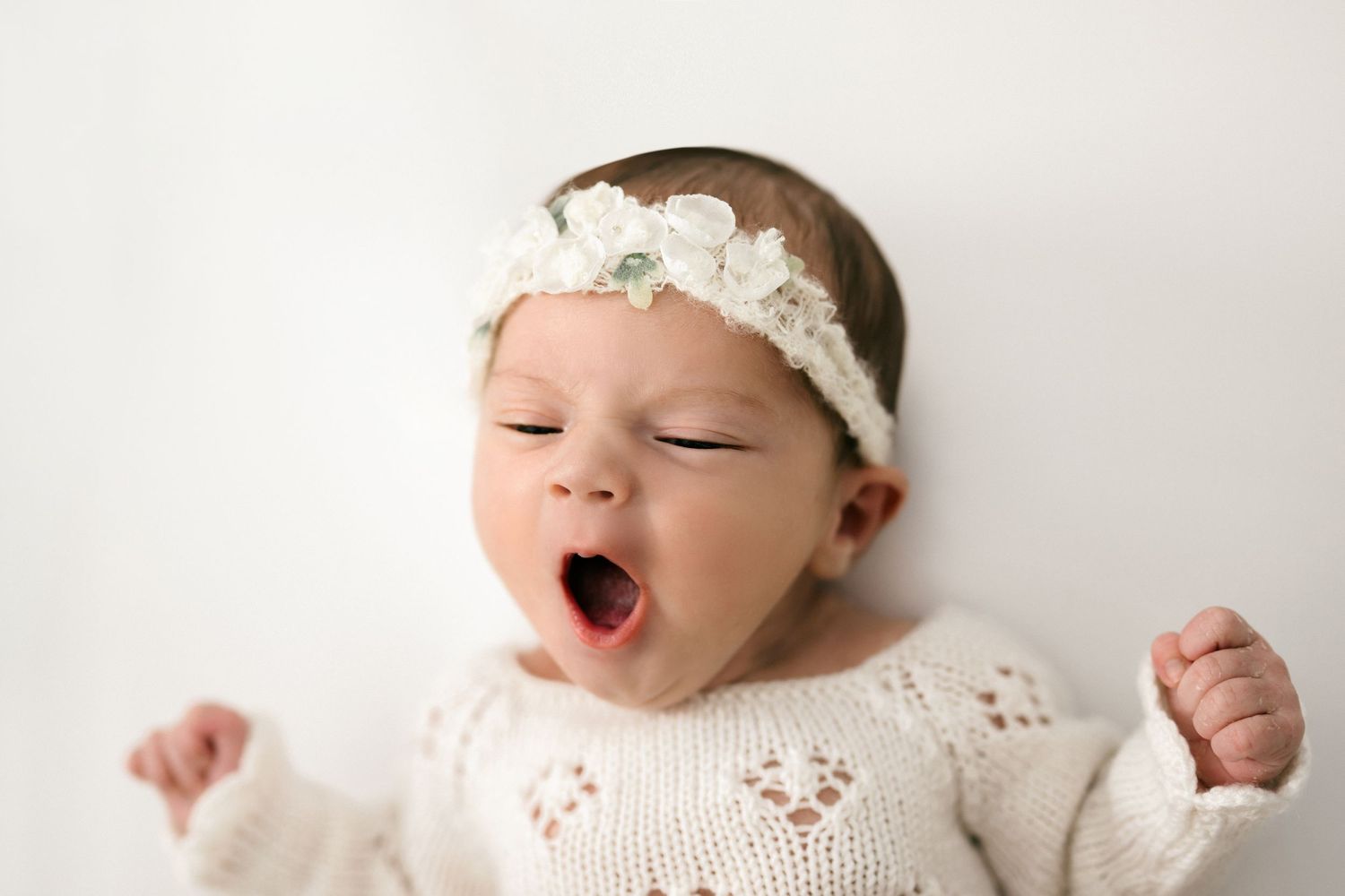 Adorable baby yawning in a white knit sweater and floral headband, lying on a soft white background.