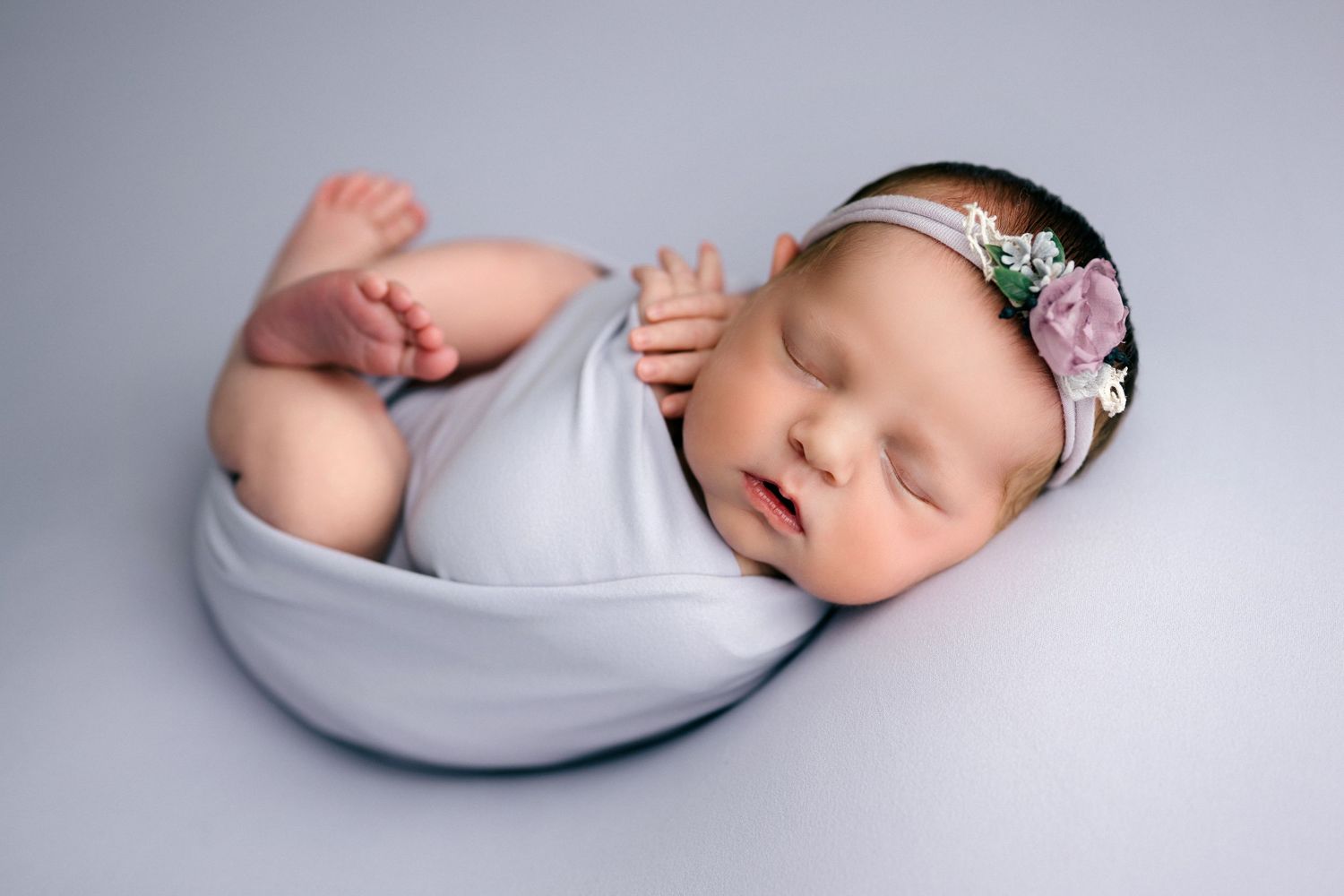 Newborn baby sleeping peacefully in a curved grey cushion wearing a delicate floral headband.