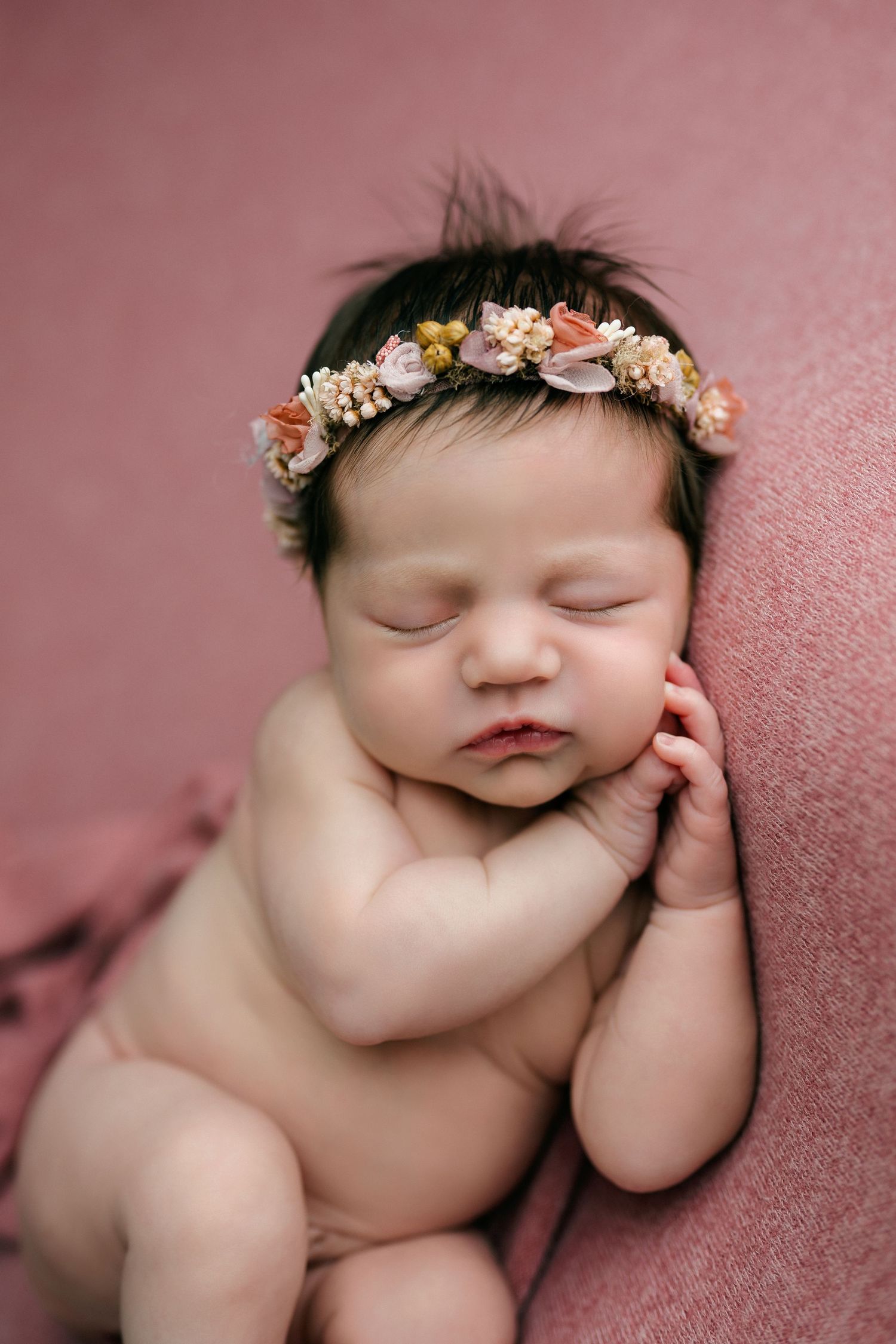 Sleeping newborn baby wearing delicate floral crown rests peacefully on pink blanket with hand near face.