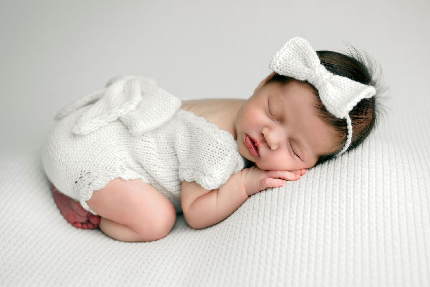 Sleeping newborn wearing white lace outfit rests peacefully on textured white blanket.