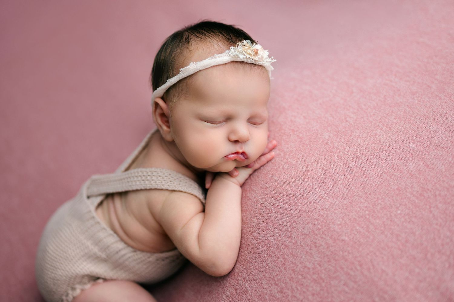 Sleeping newborn in beige outfit rests peacefully on pink blanket wearing delicate white headband.