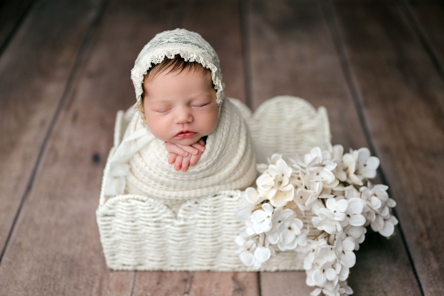 A sleeping newborn baby wearing white lace lies in a woven basket surrounded by delicate white flowers on wooden flooring.