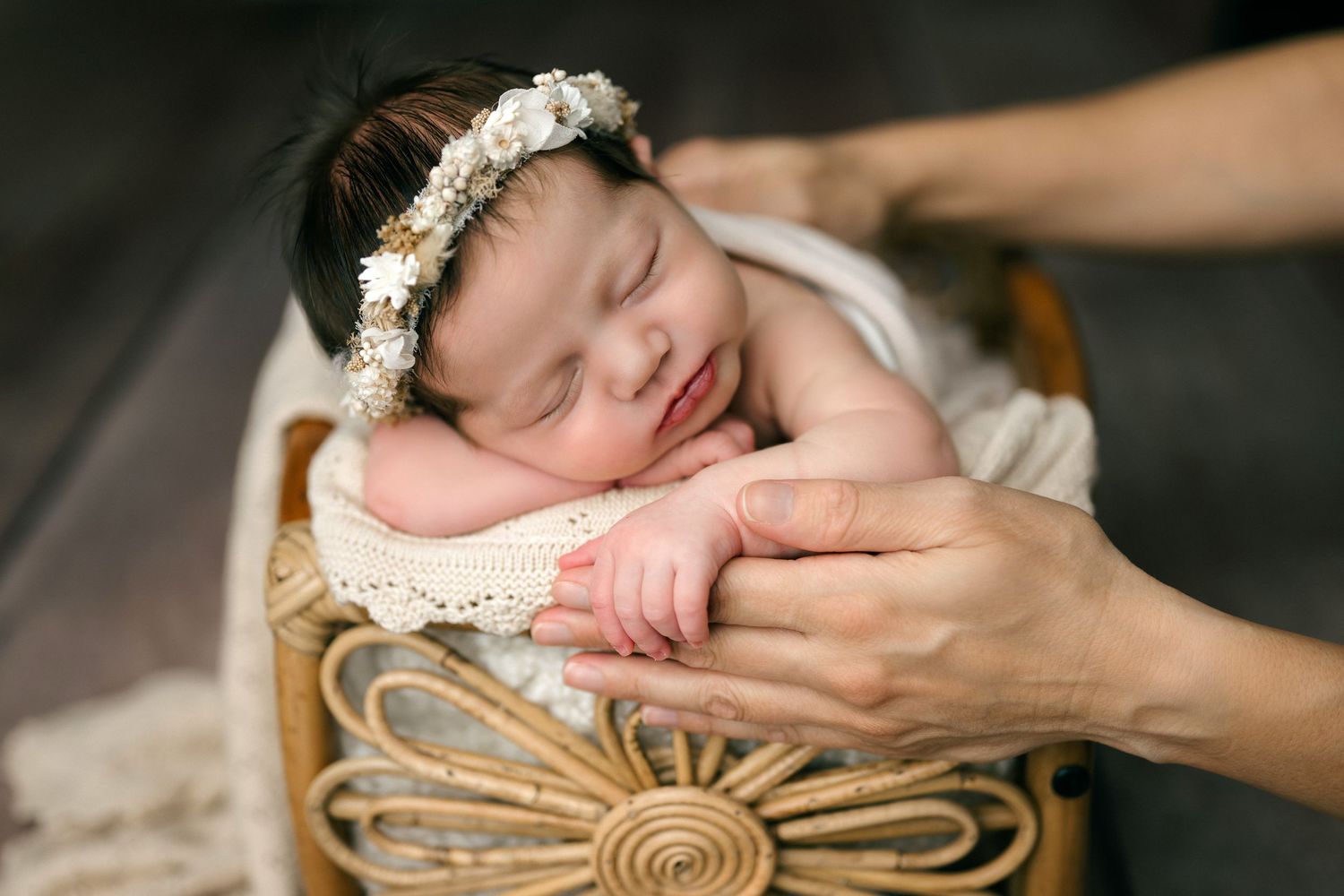 Newborn baby wearing white lace headband sleeps peacefully on brown woven basket during professional photo session.