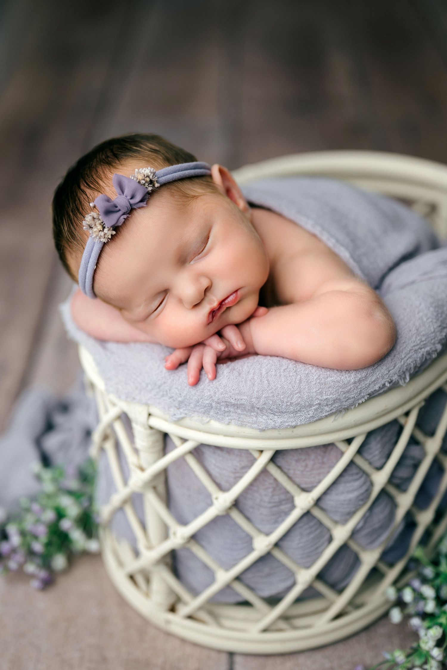 Sleeping newborn baby nestled in cream-colored woven basket with gray blanket and delicate floral accents.