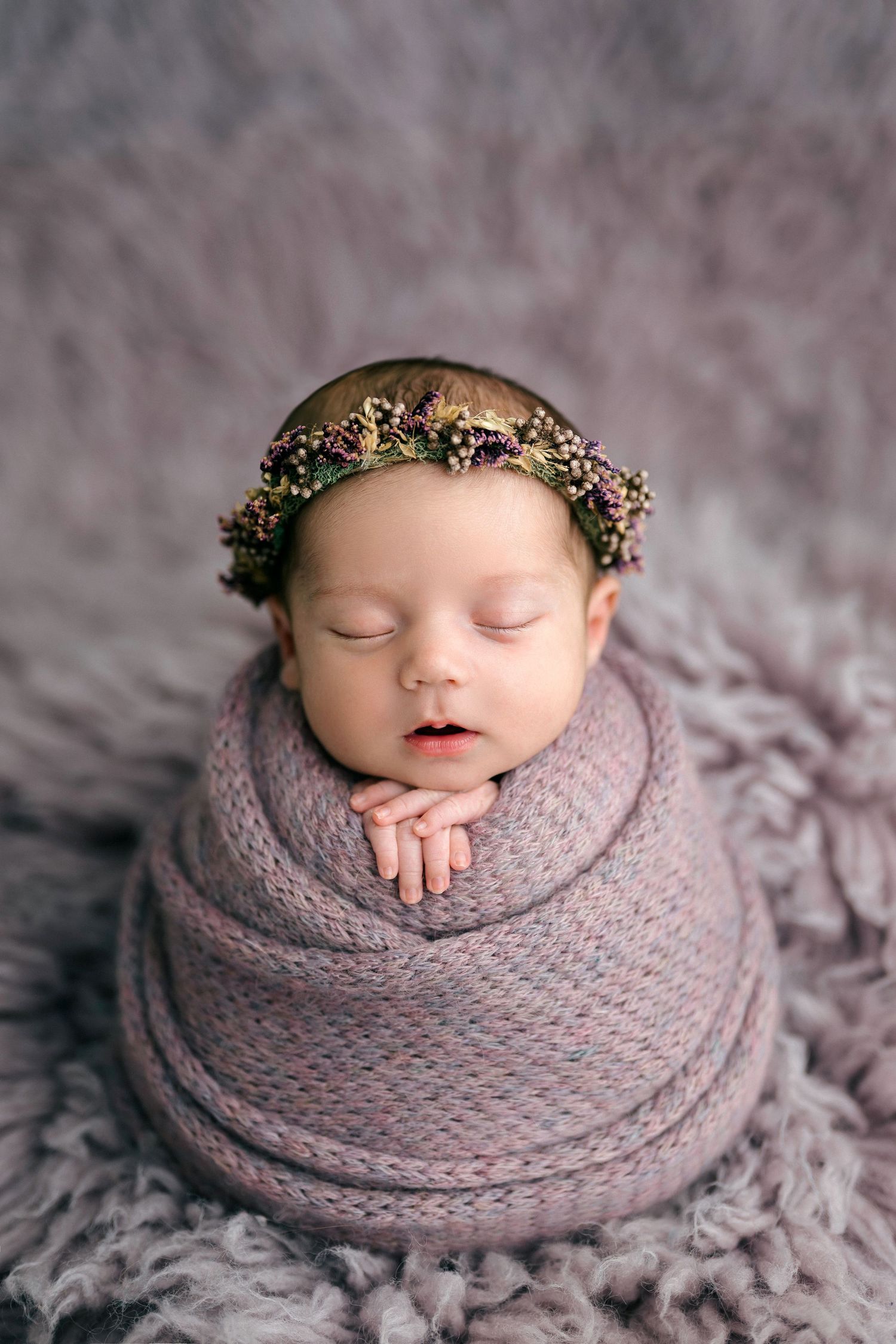 Sleeping newborn baby swaddled in pink knit wrap wearing floral crown on gray fluffy background.