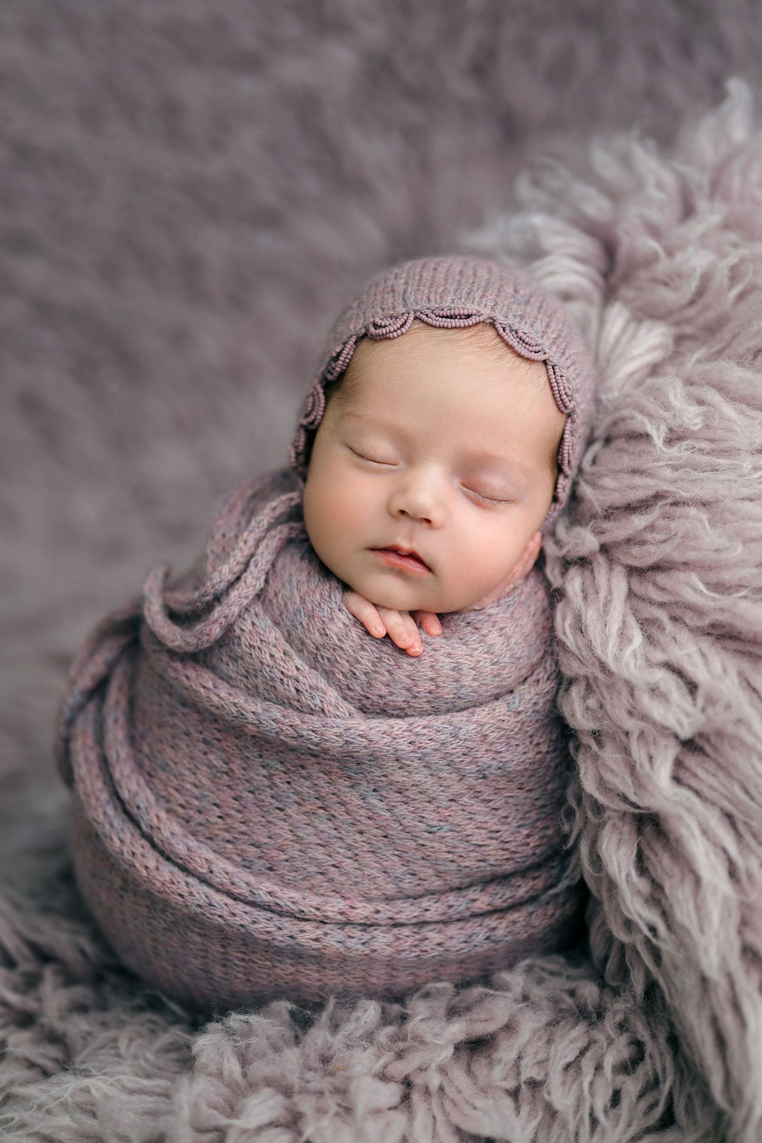 Sleeping newborn baby swaddled in a soft pink knit wrap and bonnet on a gray fluffy blanket.