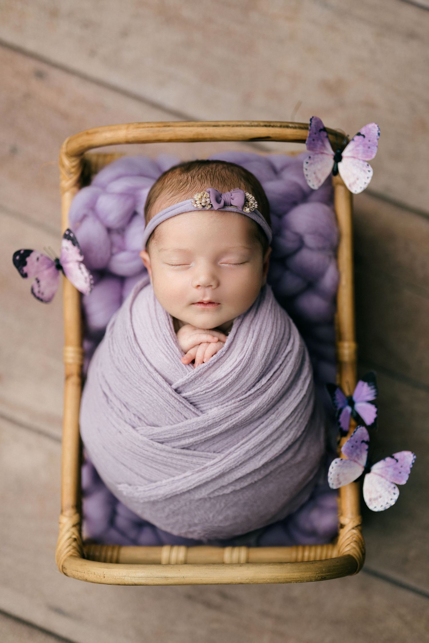 Newborn baby swaddled in purple wrap sleeping in wicker basket with butterfly decorations and lavender knit blanket.