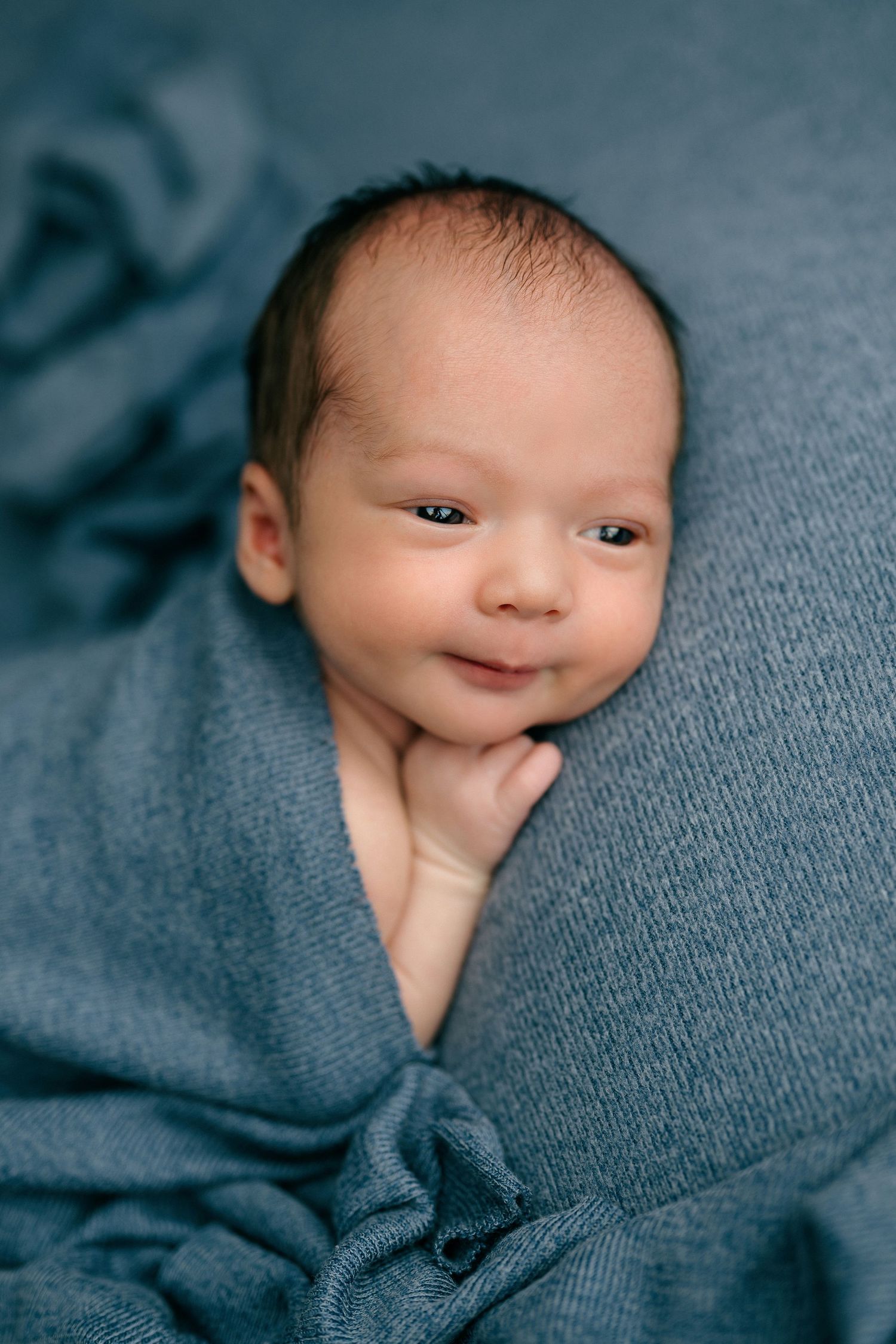 Newborn baby wrapped in a soft blue knit blanket, smiling with dark hair and bright eyes.