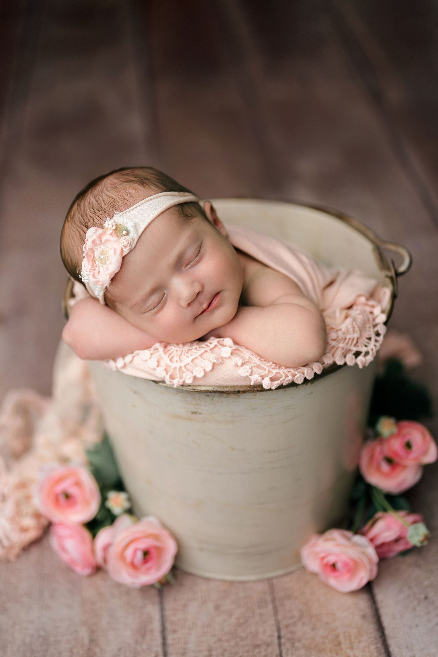 Sleeping newborn baby girl in a rustic bucket with pink lace wrap and floral headband, surrounded by pink roses.