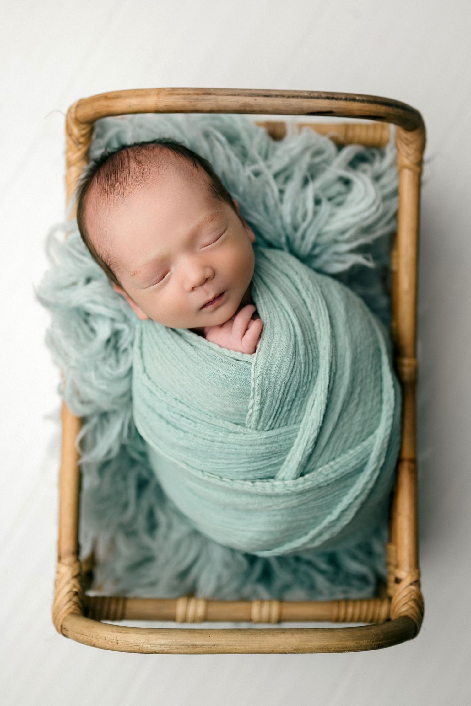 Sleeping newborn baby swaddled in a teal wrap, nestled in a small wooden basket with a soft furry blanket.