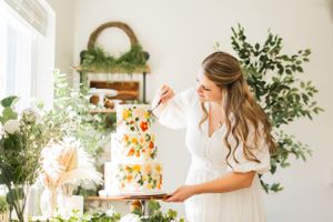 Baker decorating a tiered wedding cake adorned with colorful flowers in bright indoor setting.