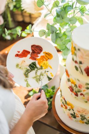 Artist mixing colorful paint on a palette next to a decorated cake with eucalyptus leaves in the background.