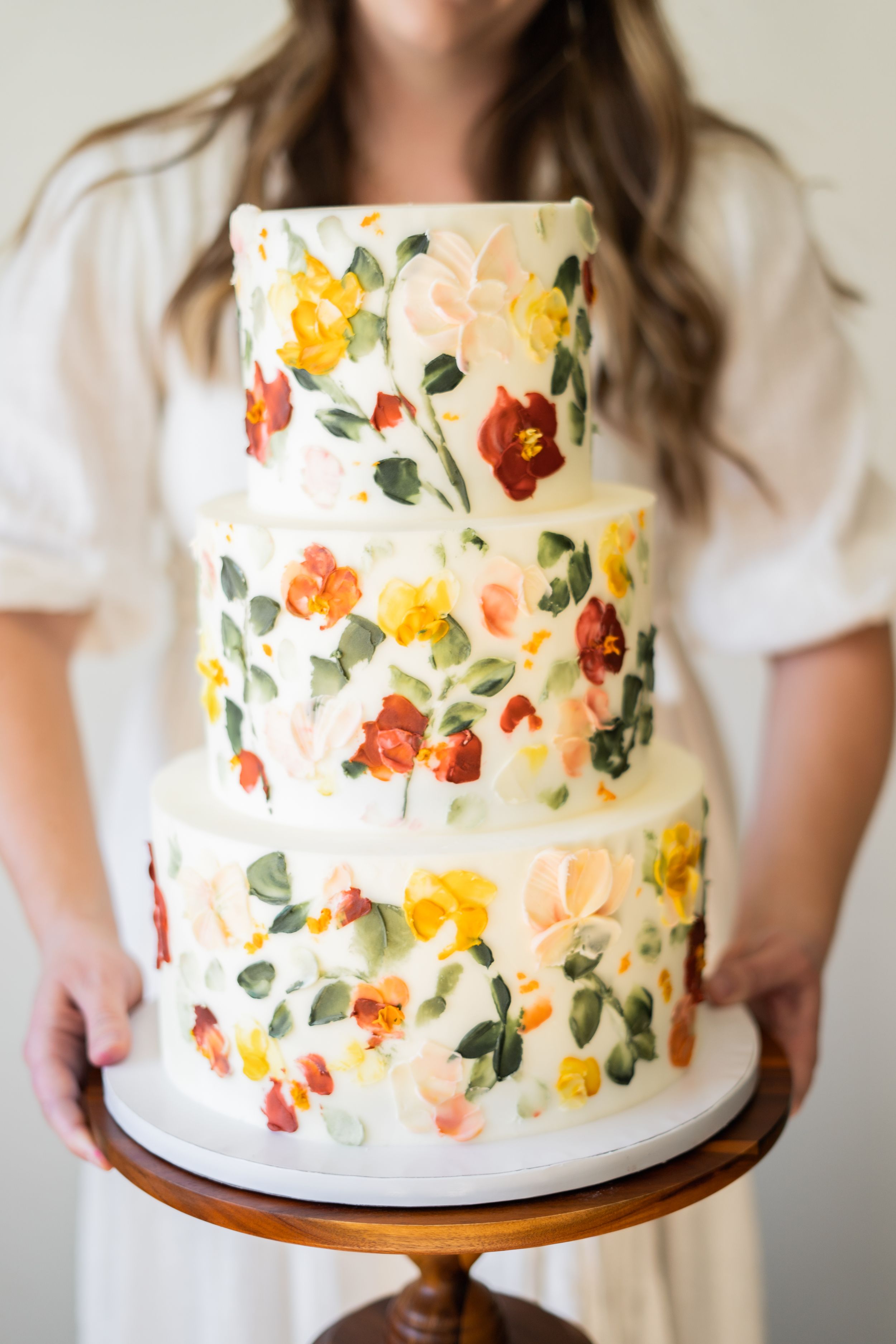 Three-tiered white wedding cake decorated with hand-painted floral designs in yellow, orange and red.