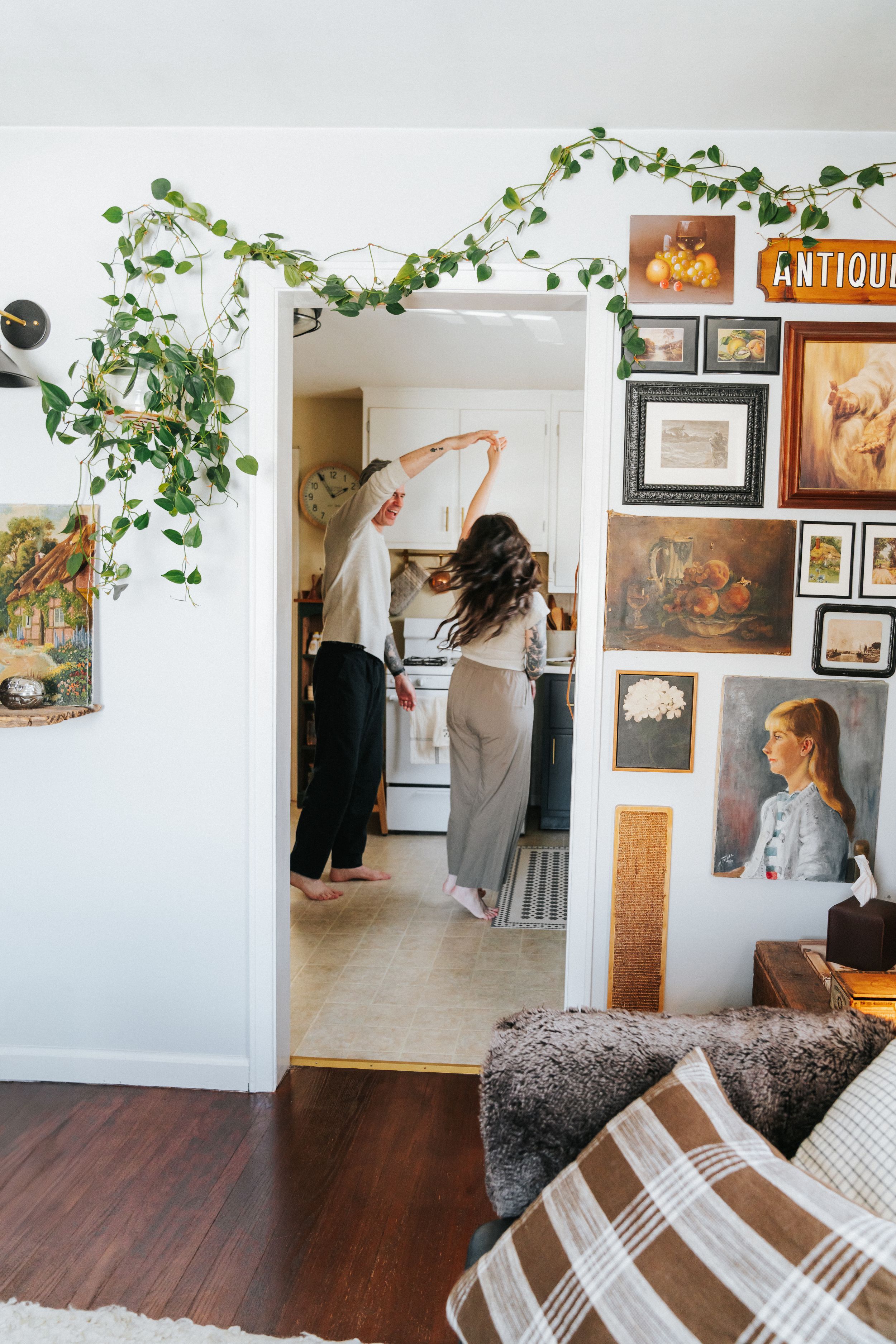Cozy home interior with climbing vines, vintage artwork gallery wall, and two people standing in a doorway.