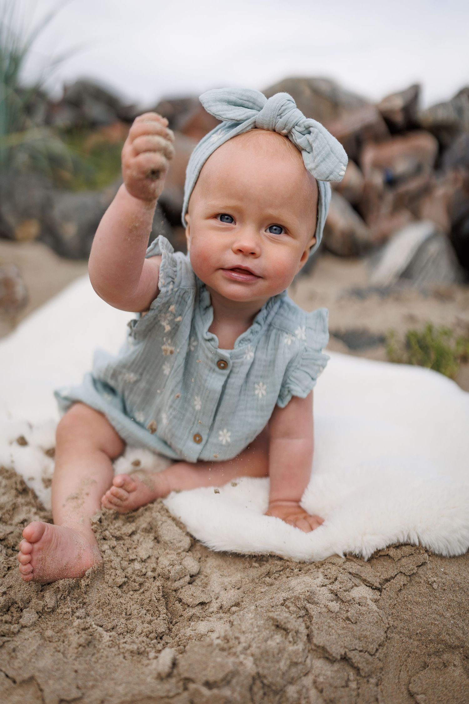 A small child wearing a light blue outfit and headband sits on sand with bare feet and raises one arm.