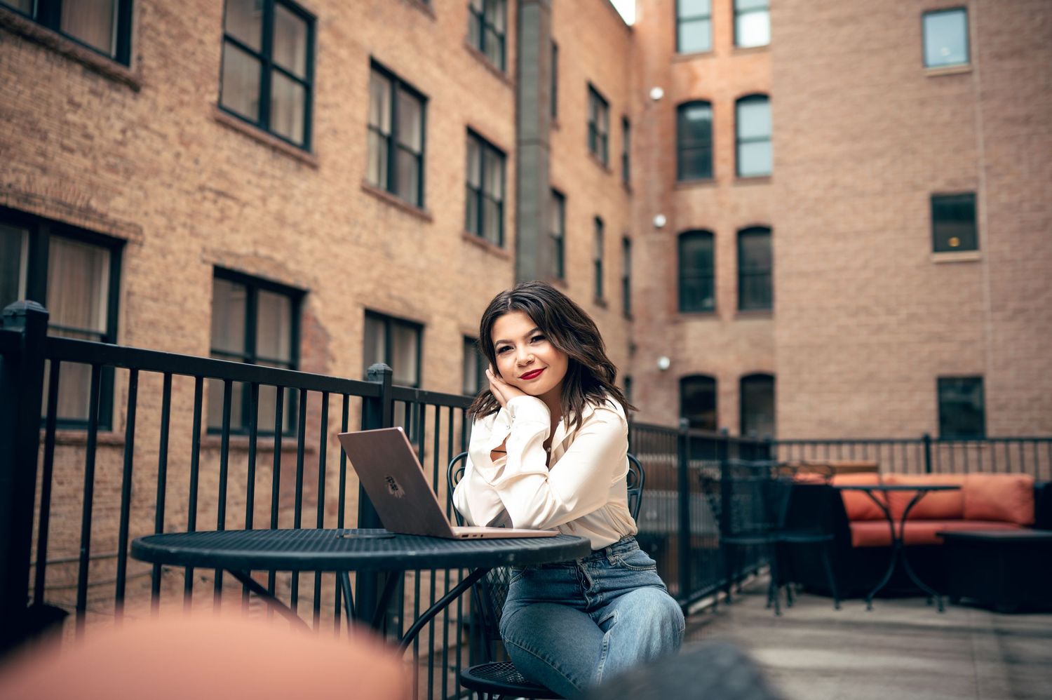 Smiling young woman with curly hair sitting outdoors with a laptop on a balcony near brick buildings.