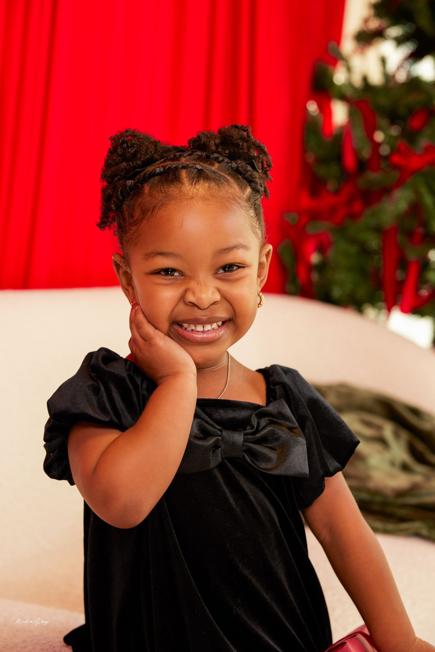 A young child in a black velvet dress smiles brightly against a red backdrop with a Christmas tree visible.