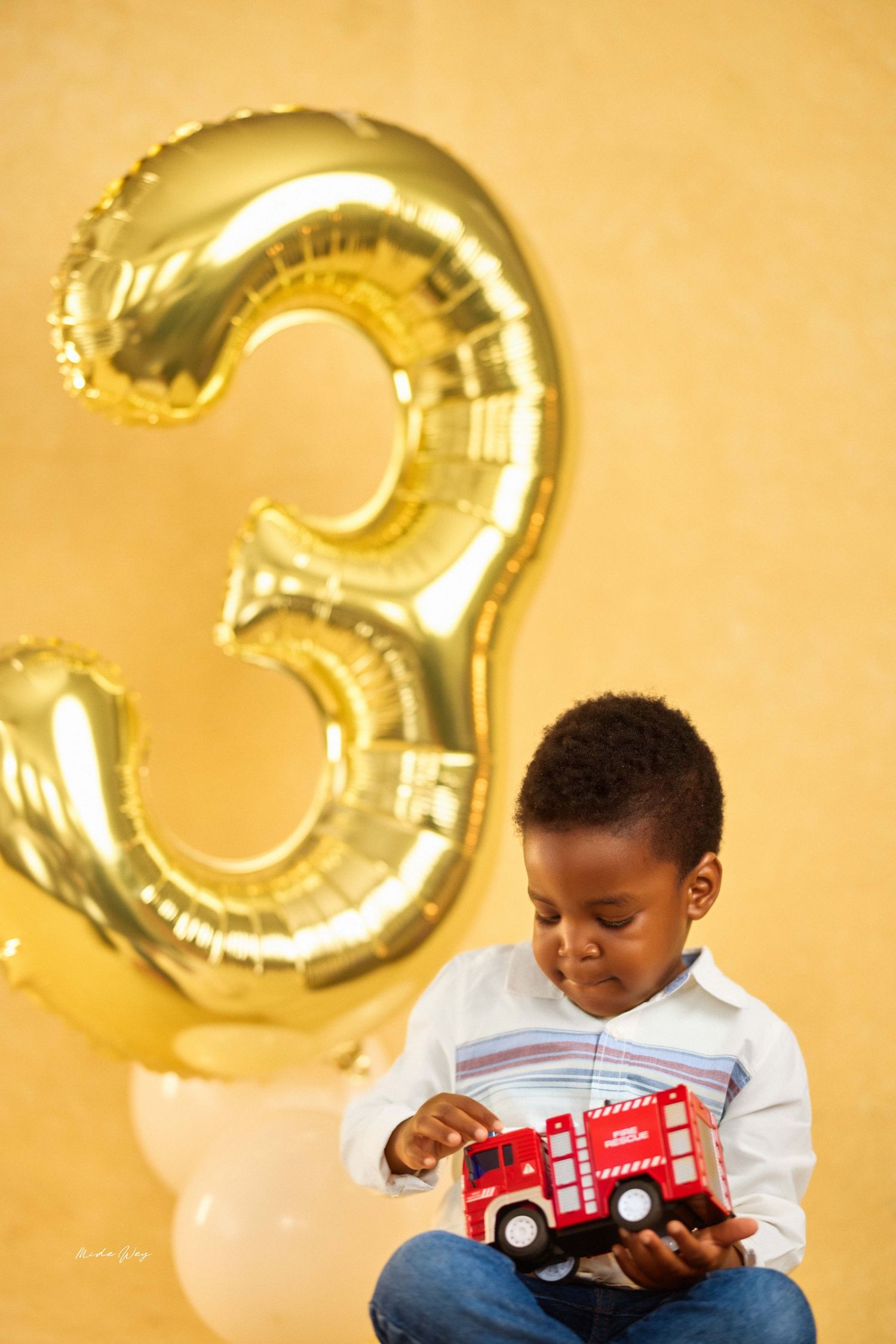 Young child in white shirt plays with red toy fire truck against golden backdrop with number 3 balloon decoration.
