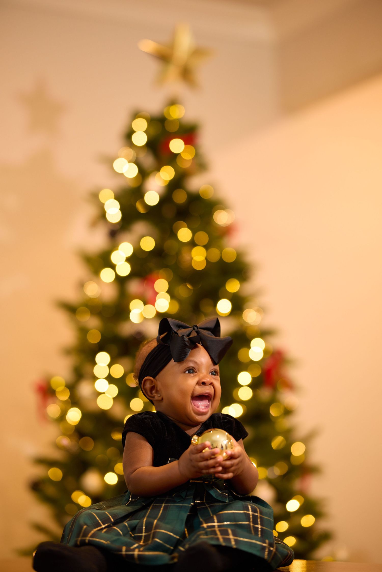 Child in plaid dress laughs joyfully while holding ornament in front of lit Christmas tree with bokeh lights and star topper.