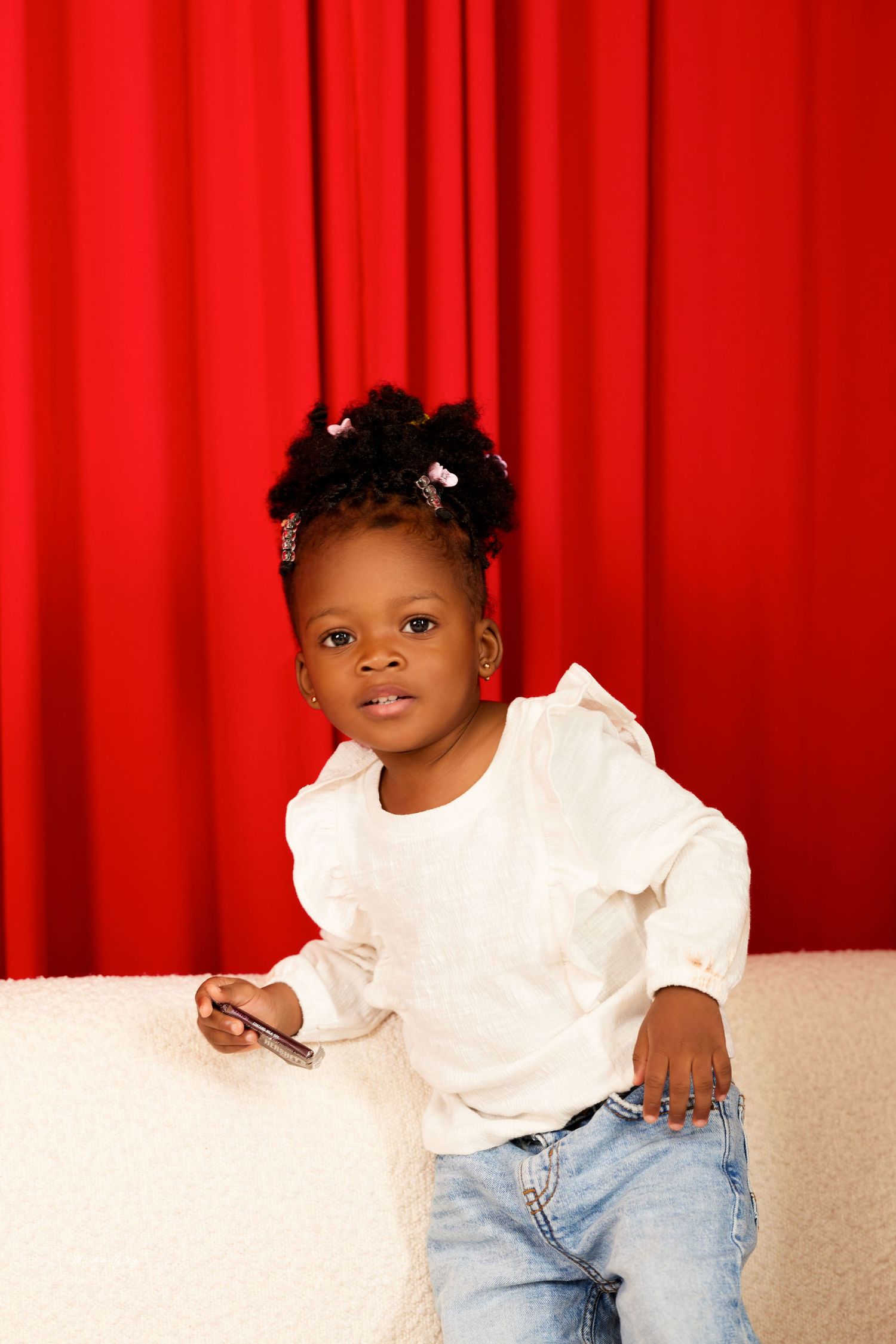 A child in a white top and jeans sits on a couch against a vibrant red curtain backdrop.