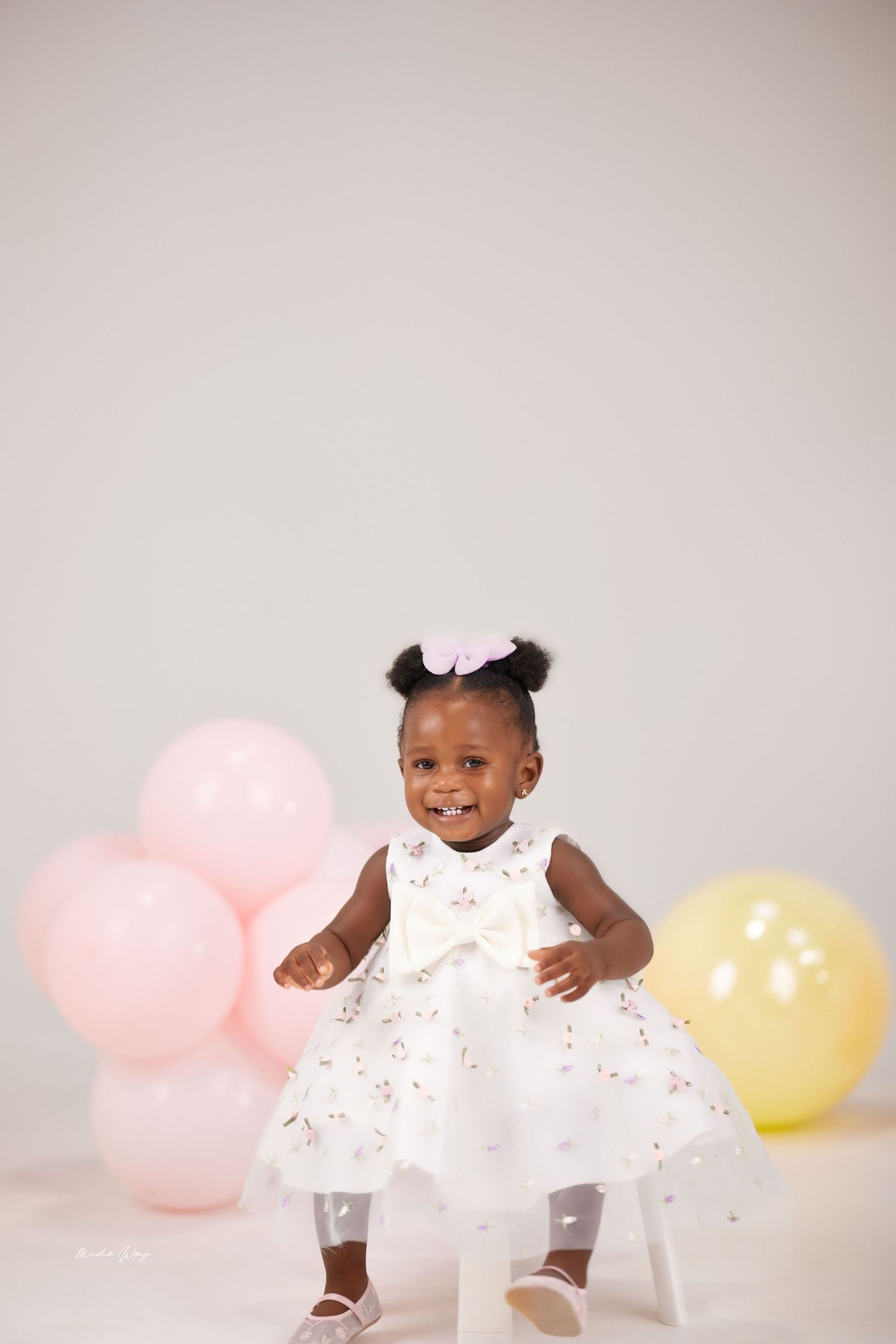 A baby in a white dress smiles while sitting and playing against a backdrop with pink and yellow balloons.