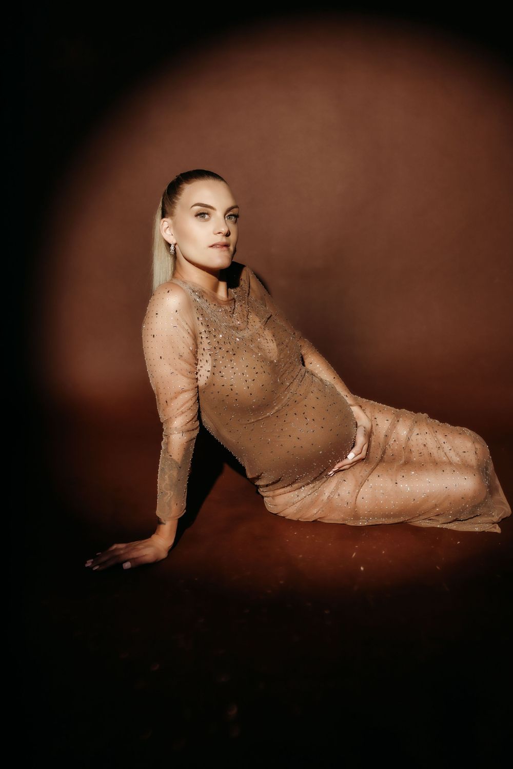 Elegant maternity portrait with sparkly bodysuit against moody brown backdrop in studio setting.