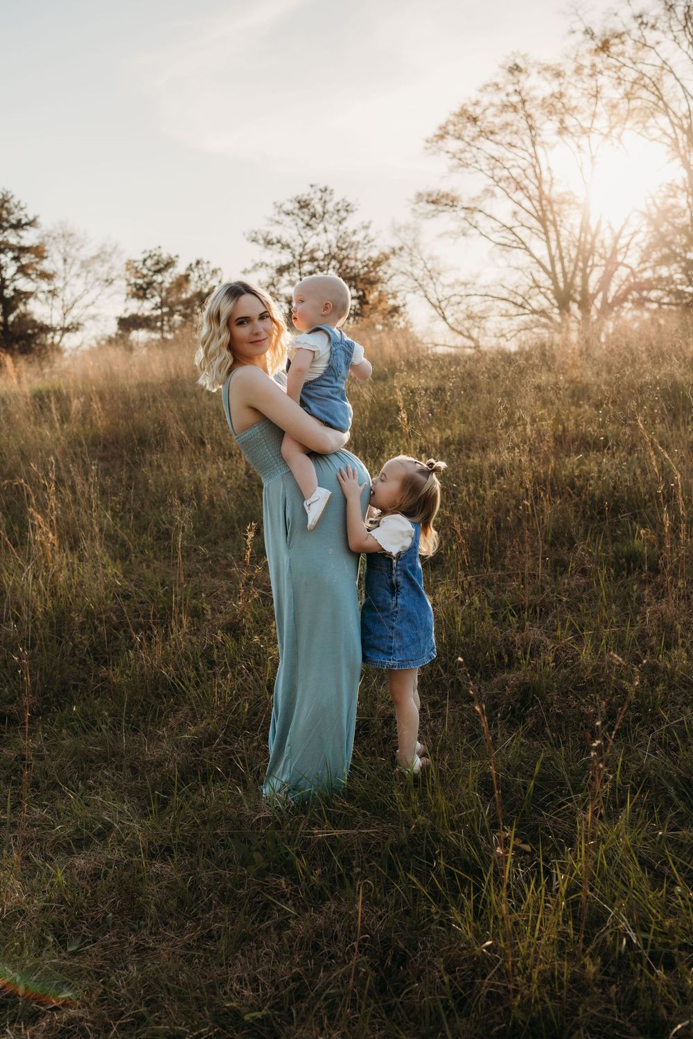Mother in blue maxi dress poses with children in field at sunset.