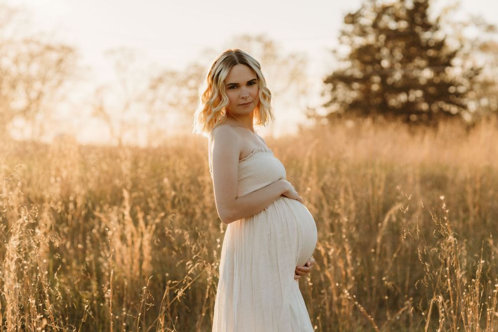 Maternity portrait of woman in white dress standing in sunlit field.
