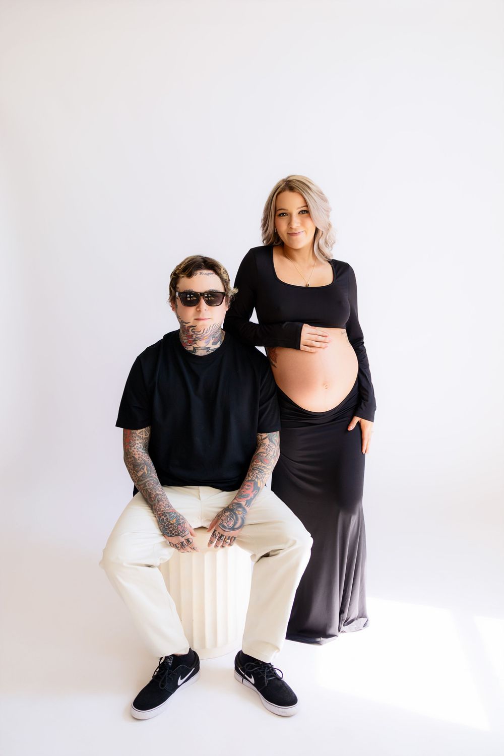 Stylish monochromatic maternity photoshoot with couple posing against white backdrop.