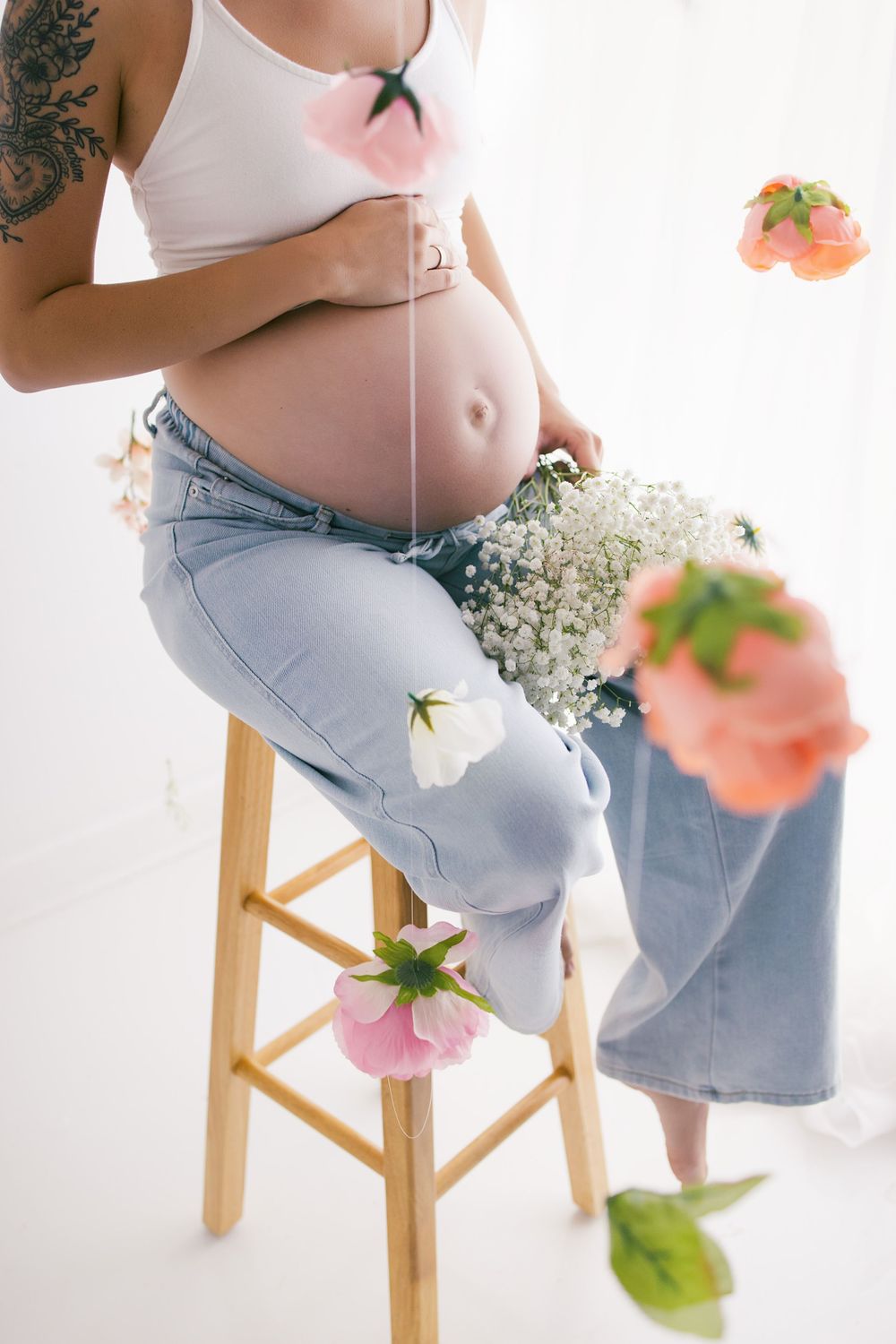 Pregnant woman in white crop top and jeans sitting on stool surrounded by hanging flowers holding a bouquet.