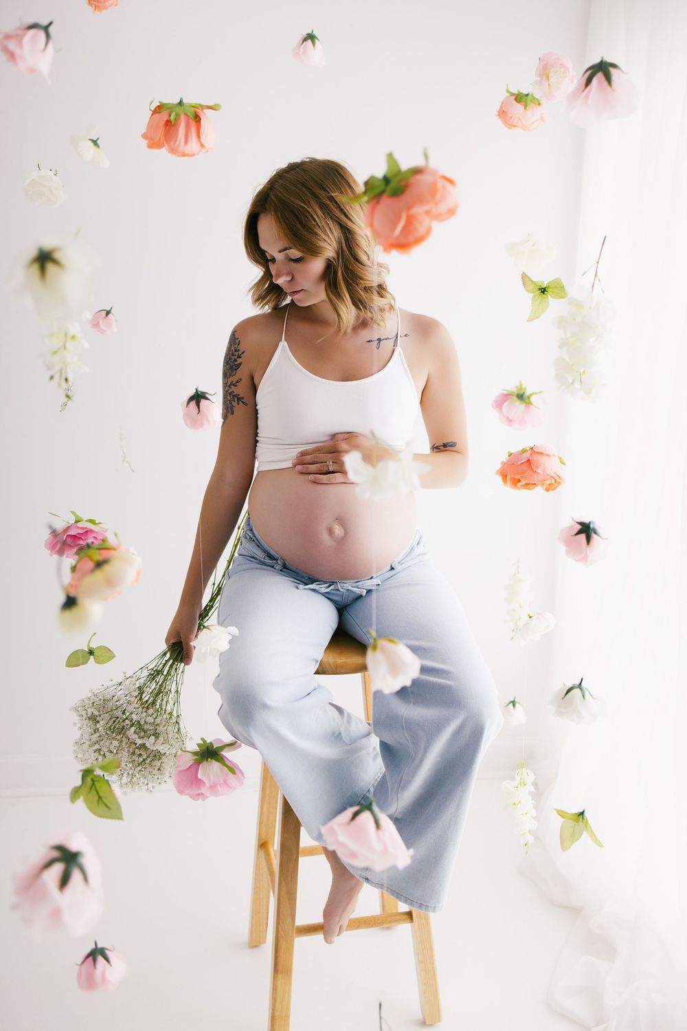 Tattooed pregnant woman in white crop top and jeans sitting on stool with floating flowers holding white blooms.