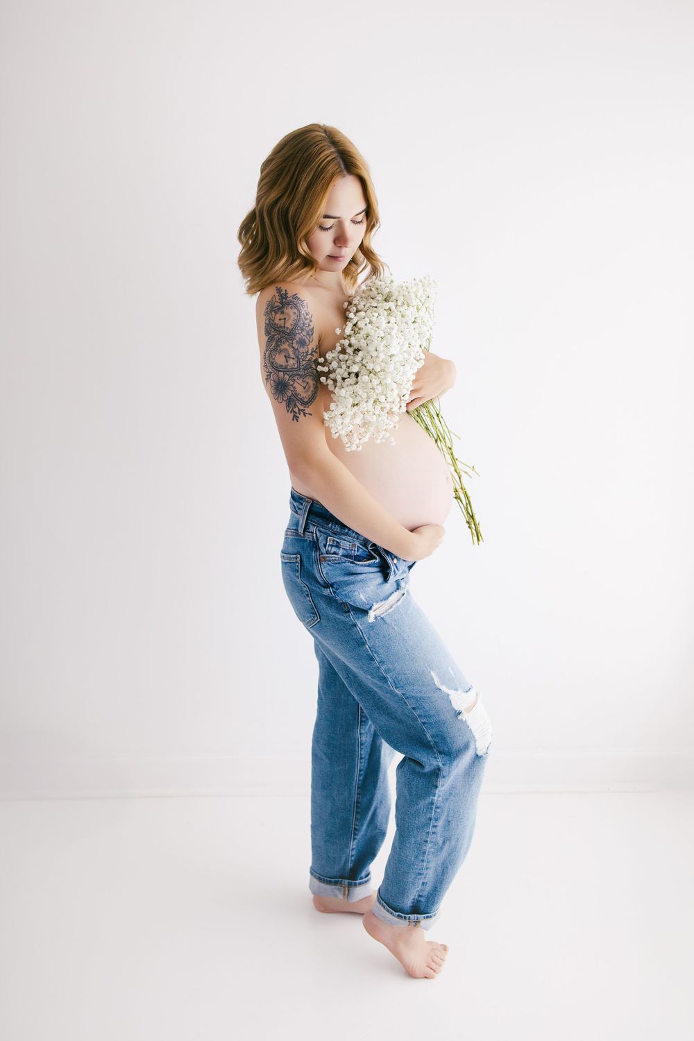 Pregnant woman in ripped jeans holding white flowers, showing baby bump and tattoo against white background.