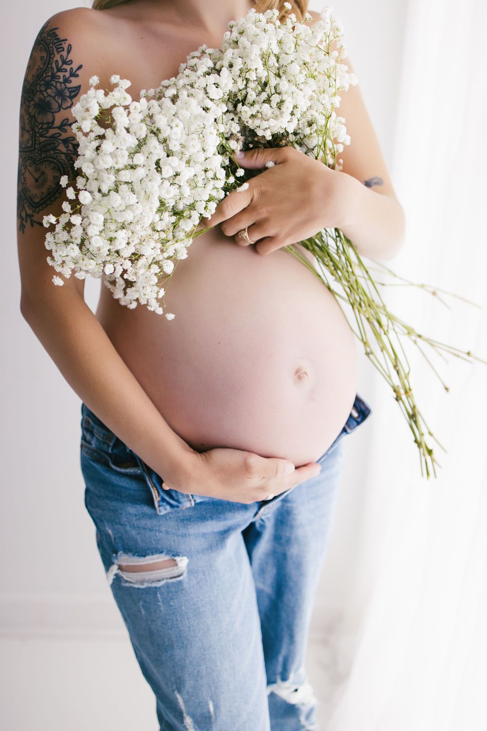 Pregnant woman in ripped jeans holding white gypsophila flowers against her bare baby bump, with a tattoo visible.