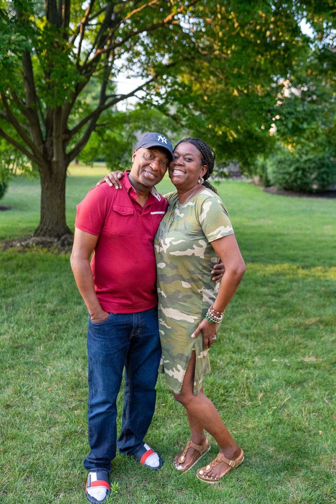 A couple poses together outdoors on a lawn with green trees in the background on a sunny summer day.