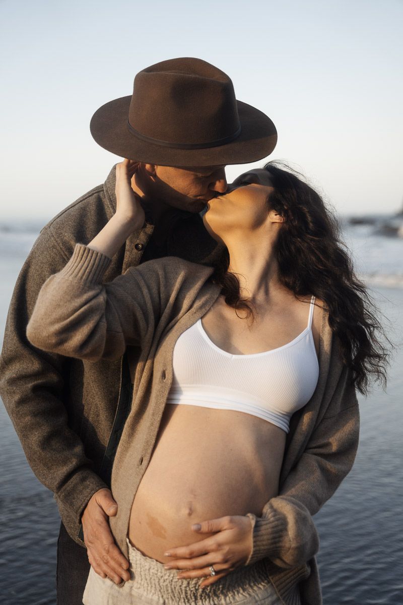Intimate maternity portrait of couple in warm earth tones sharing a romantic moment on the beach.