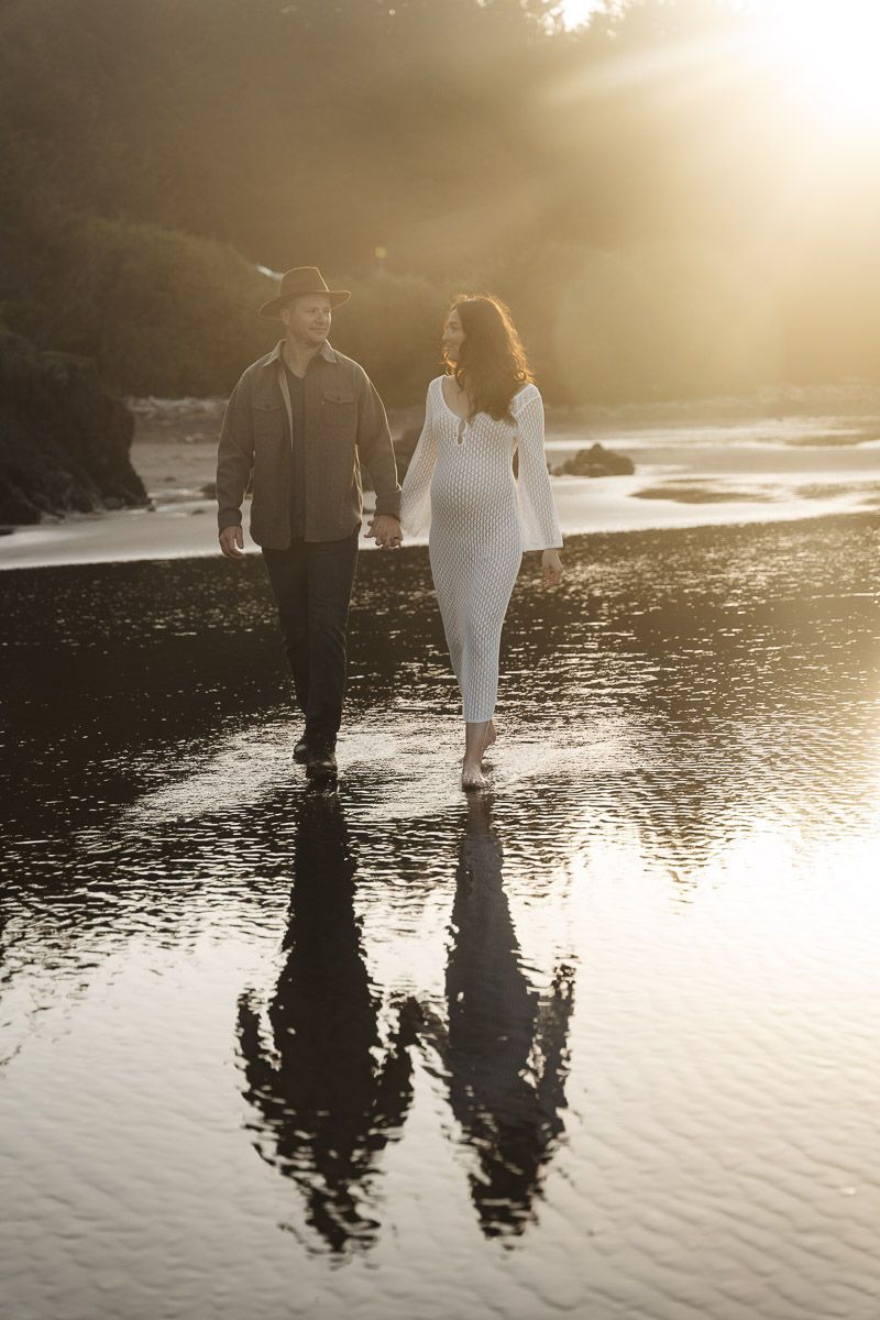 Couple walking along reflective beach at sunset with golden light casting dramatic shadows on wet sand.