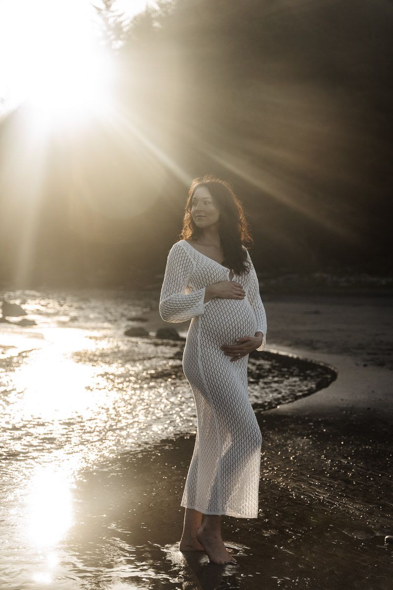 A woman in a white knit dress poses on a misty beach at sunset with dramatic light rays.