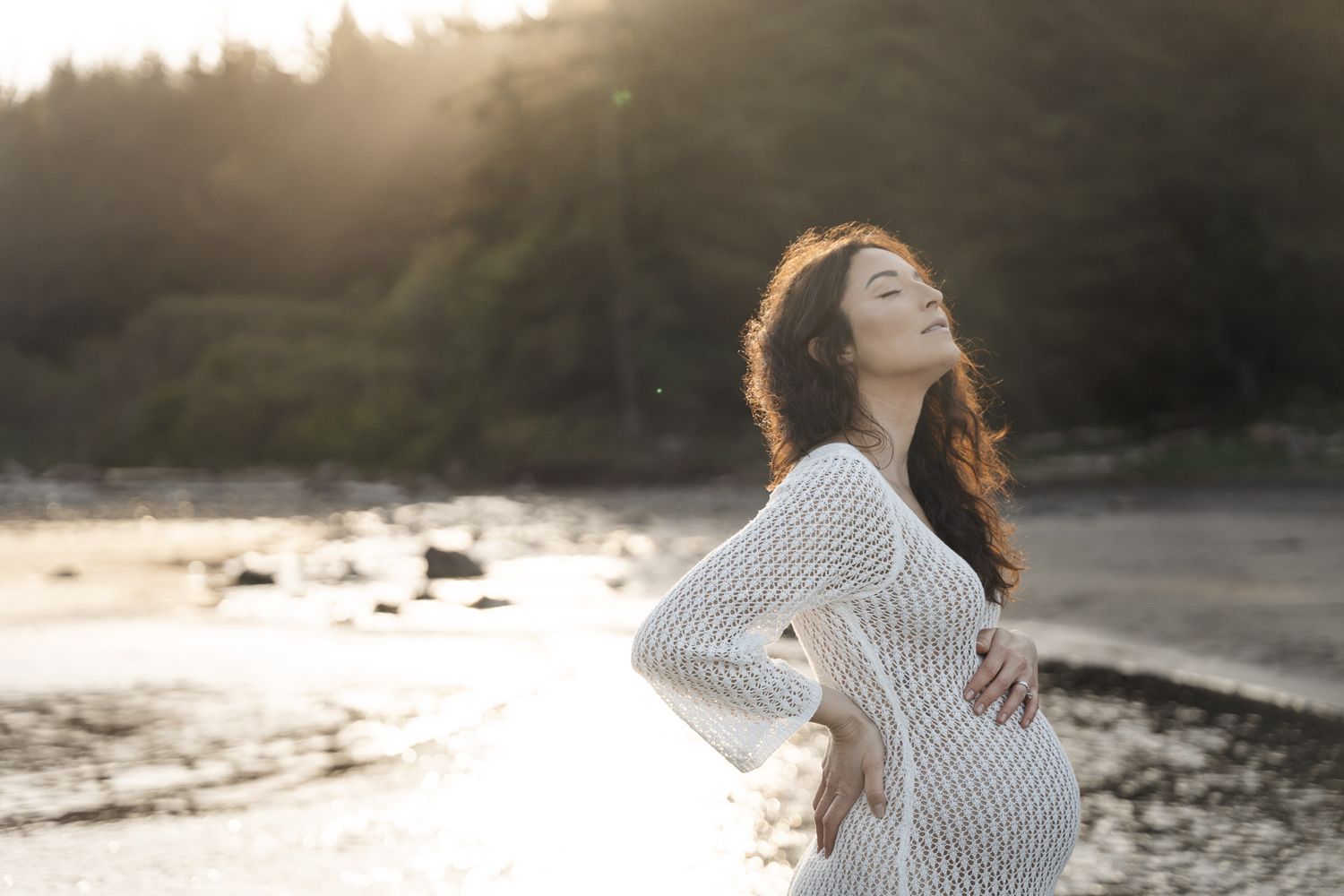 A person in a white sweater dances joyfully on a rocky beach at sunset with waves crashing in the background.