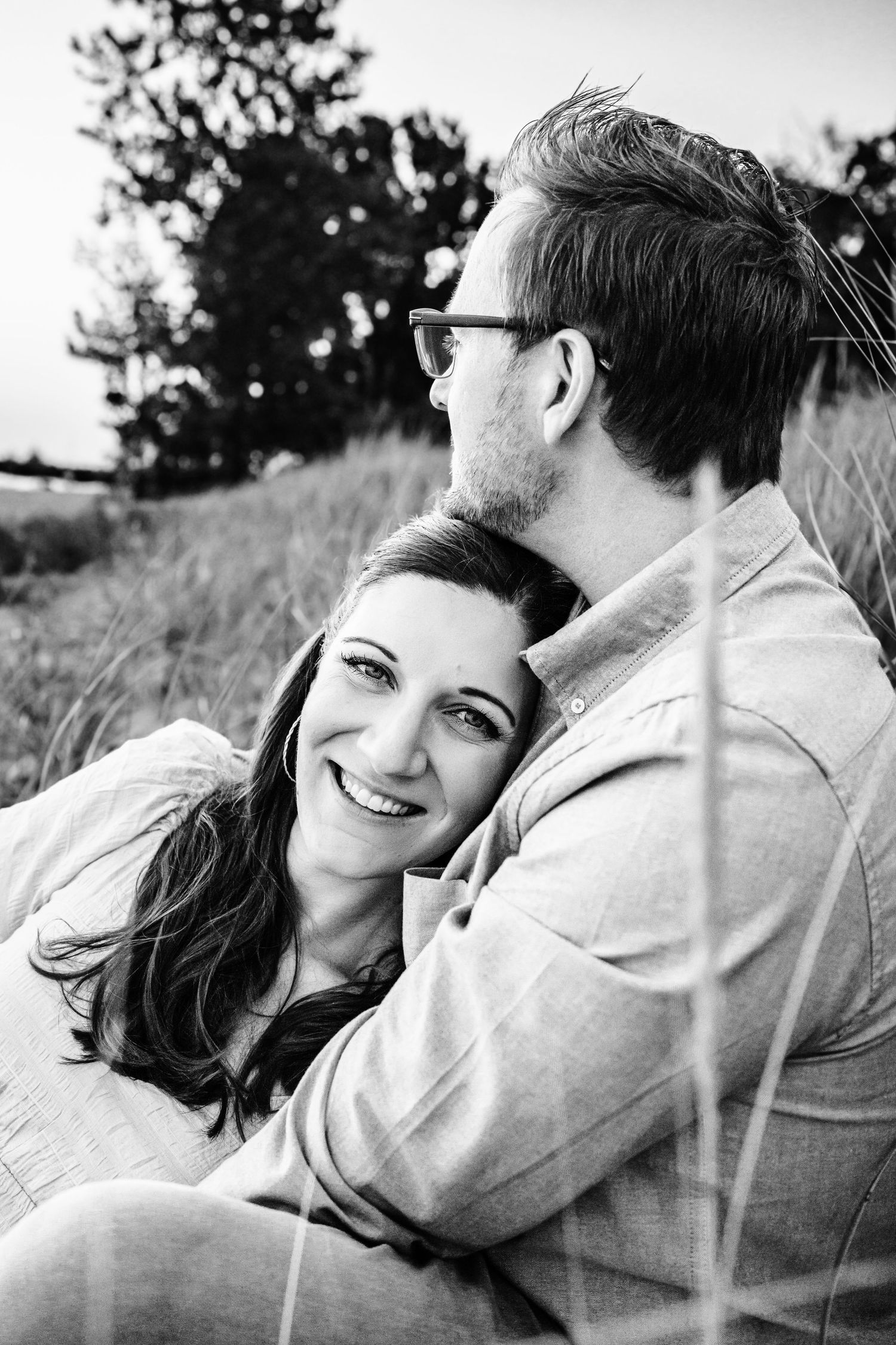 Black and white photo of a smiling woman being kissed on the head by a man wearing glasses outdoors.