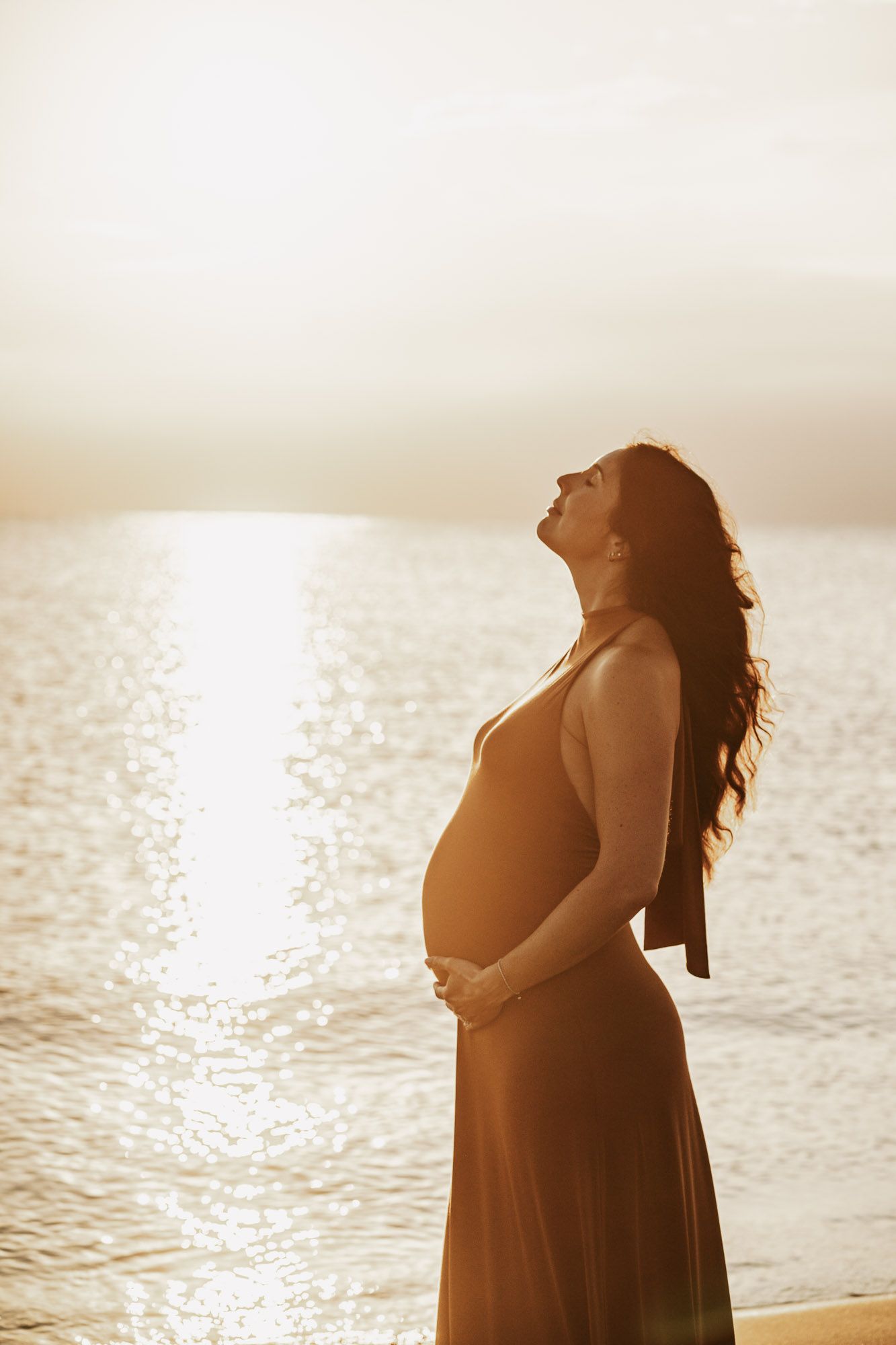 Pregnant woman in flowing dress holding her belly at golden sunset by the ocean, silhouetted against shimmering water.