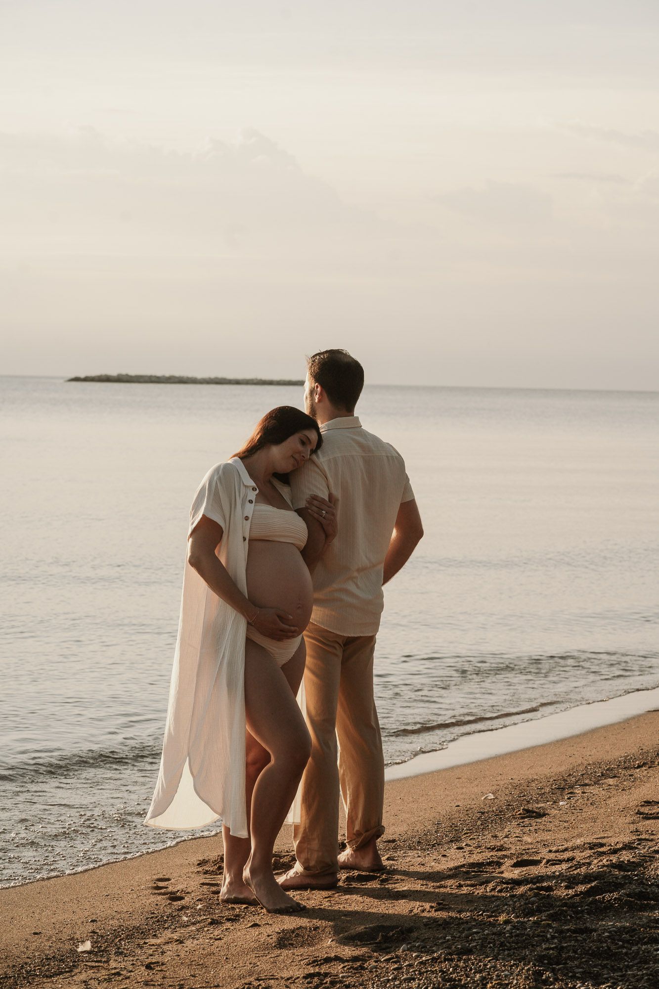 Pregnant couple standing together on a sandy beach at sunset, woman in white bikini and sheer cover-up.
