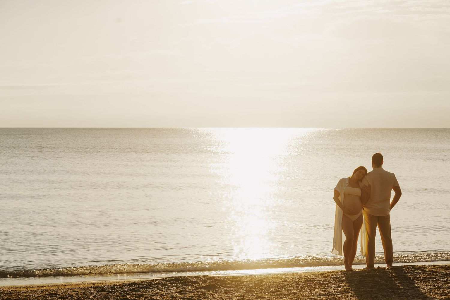 Couple standing together on a sandy beach at golden sunset, viewed from behind, overlooking calm ocean waters.