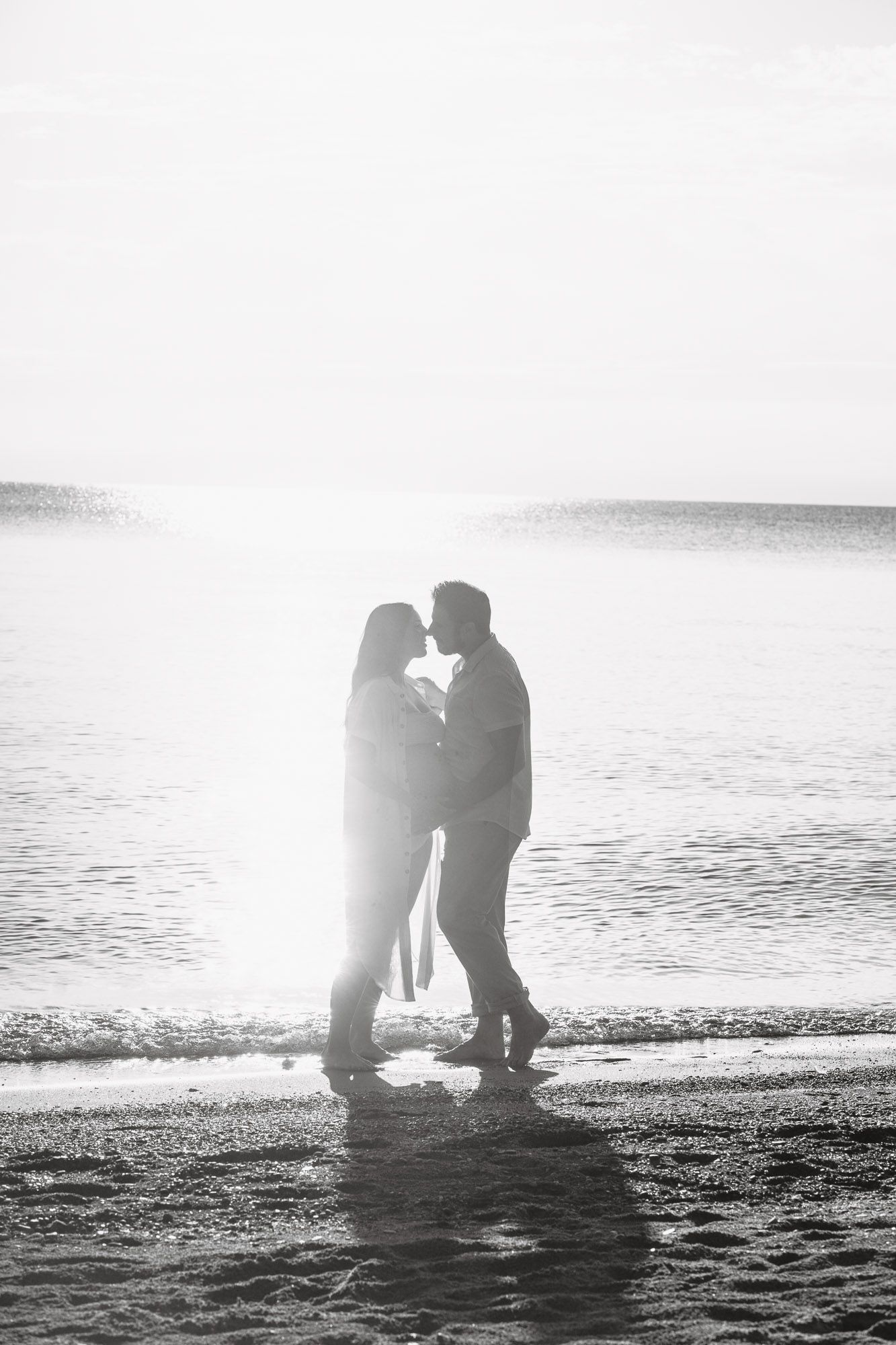 Black and white photo of a romantic couple kissing on a sunlit beach with ocean waves in the background.