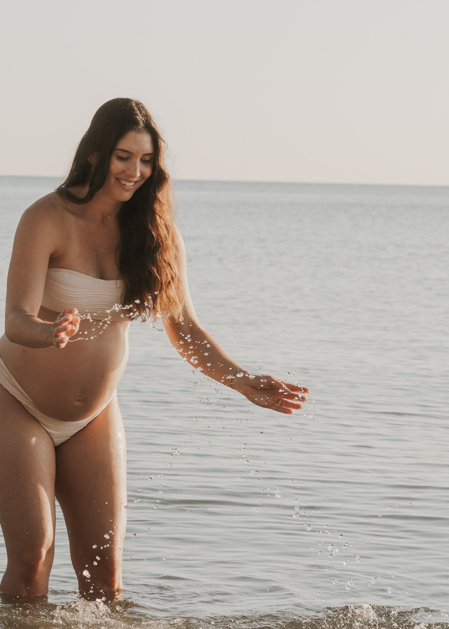 Smiling pregnant woman in beige bikini splashing water at the beach on a sunny day.